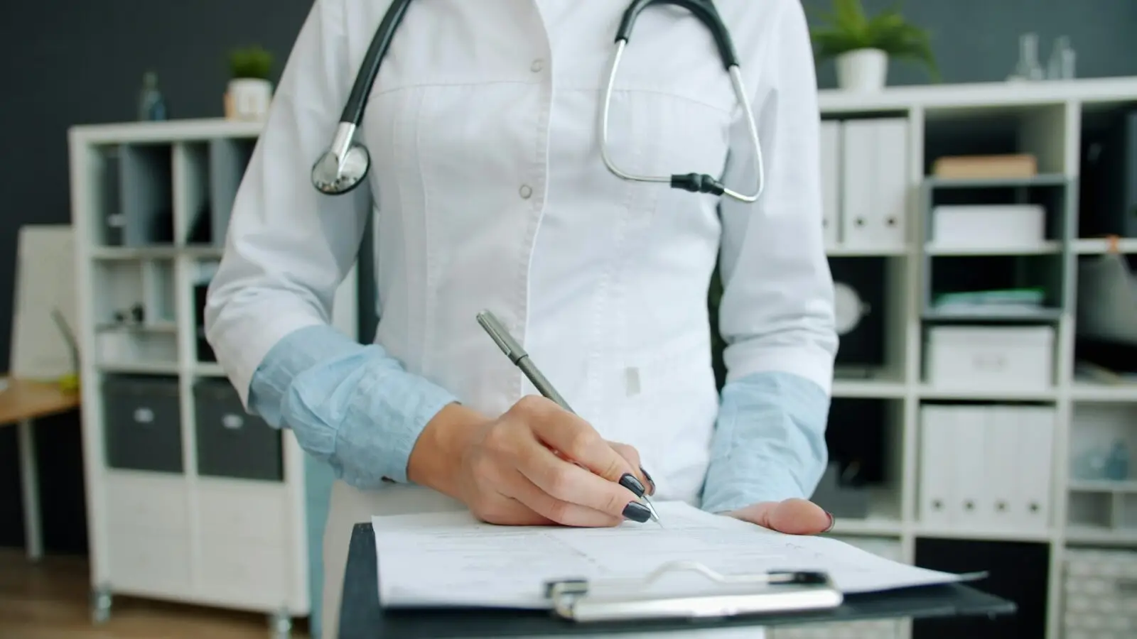 Mão de uma médica de jaleco branco preenchendo um formulário preso a uma prancheta, em um consultório